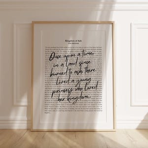 May include: A framed print with a gold-colored frame. The print features a background of small text with the words "Once upon a time" in a large, cursive font. The text "Kingdom of Ash" is at the top.