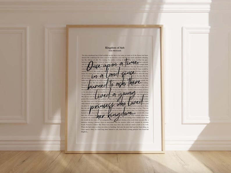 May include: A framed print with a gold-colored frame. The print features a background of small text with the words "Once upon a time" in a large, cursive font. The text "Kingdom of Ash" is at the top.