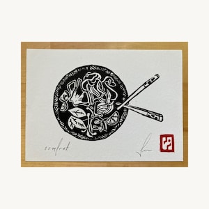 May include: A black and white linocut print on white paper depicting a bowl of noodles with chopsticks. The word comfort is written in cursive on the bottom left. A signature and a small red square stamp with a white symbol are on the bottom right.