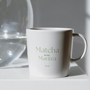 May include: A white ceramic mug with the text "Matcha is my Mantra" in a light green font. The mug has a simple design and a curved handle. The mug is on a white surface.