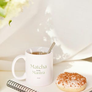 May include: A white ceramic mug with the text "Matcha is my Mantra" in green lettering, a small spoon, and a glazed donut with a crumb topping sit on a notebook. The scene is set against a soft, white background.
