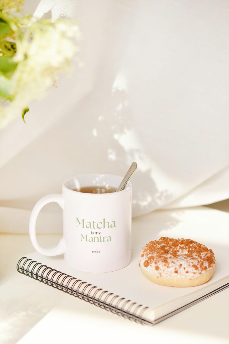 May include: A white ceramic mug with the text "Matcha is my Mantra" in green lettering, a small spoon, and a glazed donut with a crumb topping sit on a notebook. The scene is set against a soft, white background.
