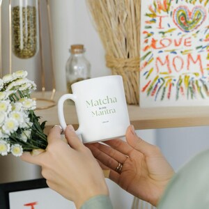 May include: A white ceramic mug with the text "Matcha is my Mantra" in green lettering. The mug is held near a bouquet of white flowers and a colorful drawing that says "I love Mom".