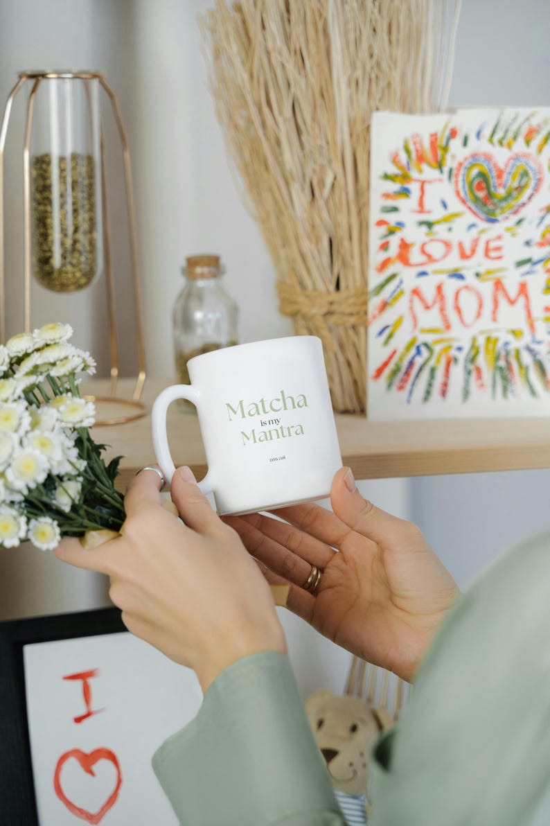 May include: A white ceramic mug with the text "Matcha is my Mantra" in green lettering. The mug is held near a bouquet of white flowers and a colorful drawing that says "I love Mom".