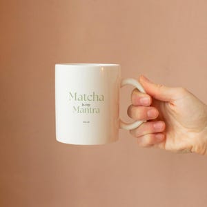 May include: White ceramic mug with the text "Matcha is my Mantra" in a light green font. The mug has a curved handle and is held against a peach-colored background. A simple, elegant design for tea lovers.