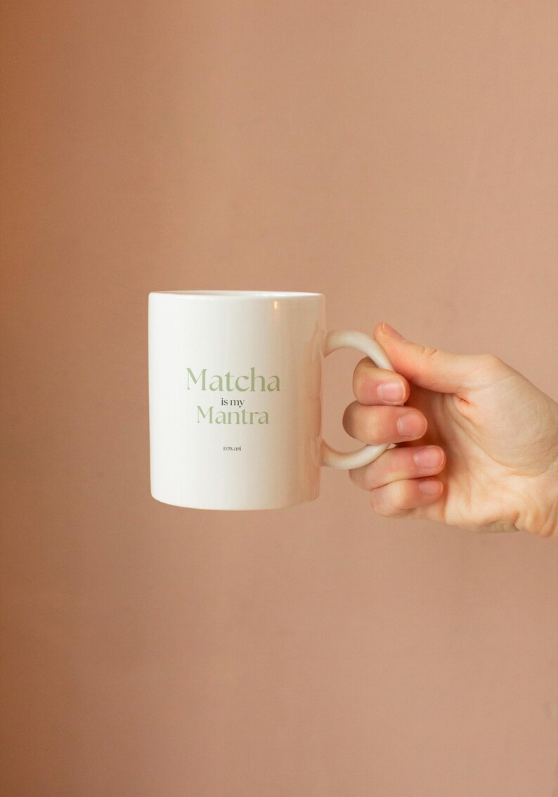 May include: White ceramic mug with the text "Matcha is my Mantra" in a light green font. The mug has a curved handle and is held against a peach-colored background. A simple, elegant design for tea lovers.