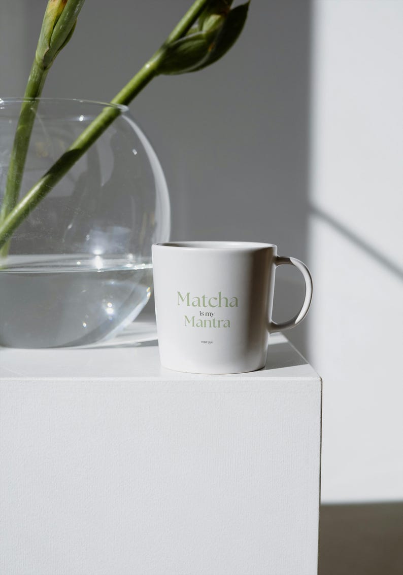 May include: A white ceramic mug with the text "Matcha is my Mantra" in a light green font. The mug has a simple design and a curved handle. The mug is on a white surface.