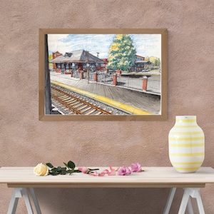 Puede incluir: Una impresión en acuarela enmarcada de una estación de tren con fachada de ladrillo rojo, un árbol y vías de tren. La obra de arte está en un marco de madera y se exhibe en una pared de color marrón rosado. Un jarrón a rayas amarillas y blancas y flores están en primer plano.