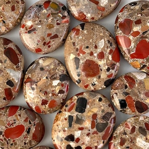 Może przedstawiać: Kolekcja owalnych kamieni o nakrapianym, mozaikowatym wyglądzie. Kamienie charakteryzują się mieszanką kolorów, w tym czerwonym, brązowym, czarnym i białym, tworząc unikalną, teksturowaną powierzchnię. Kamienie są gładkie i wypolerowane.