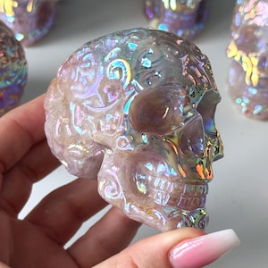 Puede incluir: Una calavera de cristal translúcida e iridiscente se sostiene en una mano, con varias otras en el fondo. La calavera tiene tallas detalladas y una superficie brillante y multicolor. Las calaveras son de color rosa claro.
