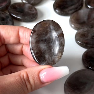 May include: A polished, oval-shaped gemstone held in a hand, with a dark brown and white marbled appearance. Several similar stones are scattered on a white surface, suggesting a collection of crystals or decorative stones.