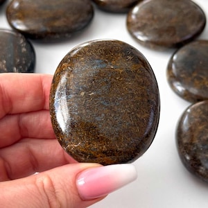 May include: A selection of polished, oval stones in shades of brown and bronze. One stone is held in a hand, displaying its smooth, reflective surface. The stones have a natural, earthy appearance.