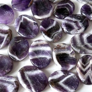 May include: A collection of polished amethyst stones. The stones are oval and rounded, with a mix of deep purple, white, and gray bands. The stones vary in size and shape, with a smooth, glossy finish.