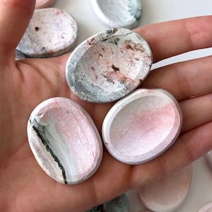 May include: A collection of oval-shaped, carved stone dishes in shades of pink, gray, and white. The dishes have a smooth, polished finish and a shallow, concave interior. Some have dark gray or black veining. The items are displayed in a hand.