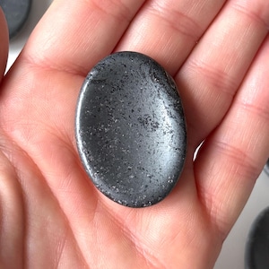 Op de afbeelding: Een gladde, ovaalvormige, donkergrijze steen met een gespikkelde textuur. De steen wordt in een open hand gehouden, met andere soortgelijke stenen zichtbaar op de achtergrond. De steen is ongeveer 5 cm lang.