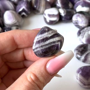 May include: A polished amethyst gemstone with white and purple bands is held in a hand. The gemstone is smooth and rounded, with a mix of dark purple and white stripes. Other amethyst stones are in the background.
