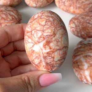 May include: Oval-shaped, polished stone with a marbled pattern of red and white. The stone is held in a hand, with several similar stones visible in the background. The stones have a smooth, glossy finish, and the red veining creates a unique, natural design.