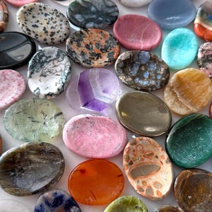 May include: A collection of polished, oval-shaped gemstones in various colors and patterns. The stones include shades of pink, green, orange, blue, and brown, with unique veining and speckled textures. The gemstones are arranged closely together, filling the frame.