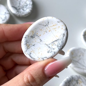 May include: White Howlite oval-shaped stones with gray and gold veining. The stones have a polished finish and are held in a hand. Other stones are visible in the background.