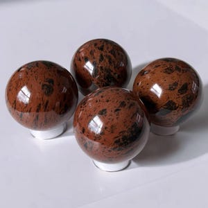 May include: Four polished obsidian spheres with a reddish-brown hue and black speckling. Each sphere sits atop a small, white base. The spheres are arranged closely together, reflecting light.