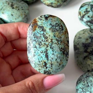 Puede incluir: Una piedra de turquesa pulida de forma ovalada con un color base vibrante en cian y azul claro, con intrincadas vetas de matriz de color marrón oscuro a negro en toda la superficie. La piedra tiene un acabado liso y brillante.