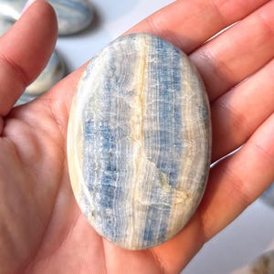 May include: An oval-shaped polished stone with a blue and cream banded pattern. The stone is held in a hand, showcasing its smooth texture and natural color variations. The stone's surface has a subtle sheen.