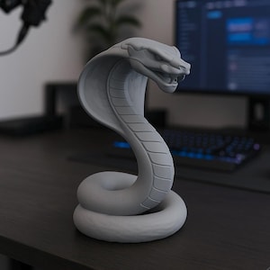 Puede incluir: Una estatua de cobra gris con una superficie detallada y texturizada. La serpiente está enrollada y erguida, con su capucha extendida y la boca ligeramente abierta. La escultura está sobre una superficie oscura, con un ordenador y un teclado al fondo.