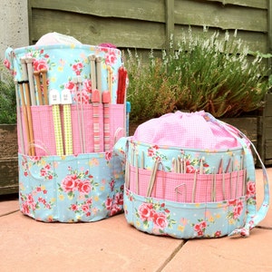 May include: Two floral patterned fabric storage containers for knitting needles. The larger container is cylindrical and has a pink and blue floral pattern. The smaller container is round and has a pink and blue floral pattern with a pink lining. Both containers are filled with knitting needles.