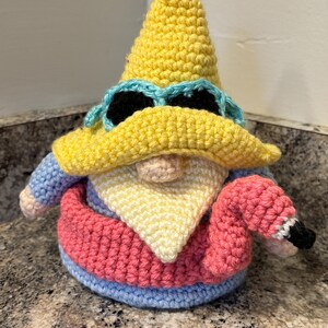 May include: A crocheted gnome figure wearing a yellow pointed hat with light blue sunglasses, a light blue shirt, and a pink inner tube. The gnome has a pale peach nose and a cream-colored beard, and it is holding a small pink flamingo floatie.