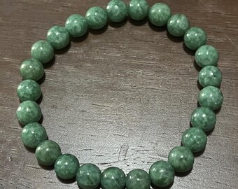 May include: A green beaded bracelet. The bracelet features round, polished beads in various shades of green. The beads are strung together, forming a circular shape, designed for the wrist.