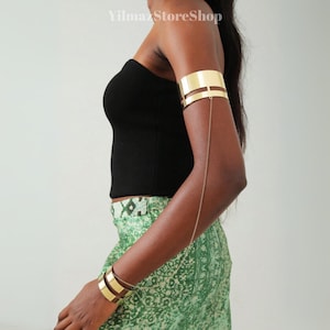 Pode incluir: Punho de braço e pulseiras douradas. O punho do braço apresenta um design de faixa dupla com uma corrente delicada que se estende até o ombro. As pulseiras são de cor dourada combinando. O texto "YilmazStoreShop" é visível.