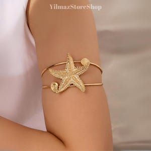 May include: Gold-toned arm cuff featuring a starfish and seashell design. The cuff has a double-banded structure with a starfish in the center and a seashell on the upper band. The cuff is a beach-themed accessory.