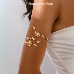 May include: Gold-coloured arm cuff with floral and leaf designs. The cuff wraps around the upper arm with two bands, adorned with flower and leaf details. The jewellery is embellished with small, sparkling accents.