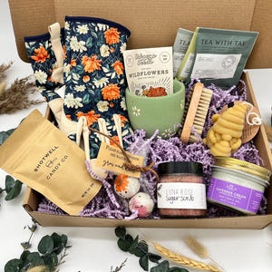 May include: A gift box filled with self-care items. Includes floral gardening gloves, a green ceramic cup, tea packets, a wooden nail brush, a honey-shaped soap, a sugar scrub, a lavender cream jar, and Shotwell Candy Co. candy.