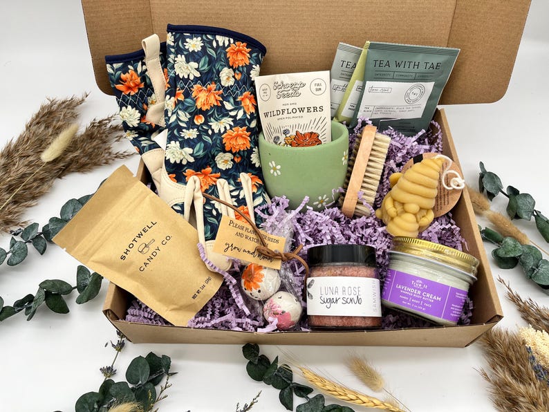 May include: A gift box filled with self-care items. Includes floral gardening gloves, a green ceramic cup, tea packets, a wooden nail brush, a honey-shaped soap, a sugar scrub, a lavender cream jar, and Shotwell Candy Co. candy.