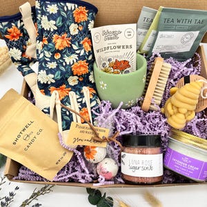 May include: A gift box filled with gardening and self-care items. Includes floral gardening gloves, wildflower seeds, tea, a sugar scrub, lavender cream, a bee-shaped candle, and candy. The box is lined with purple paper.