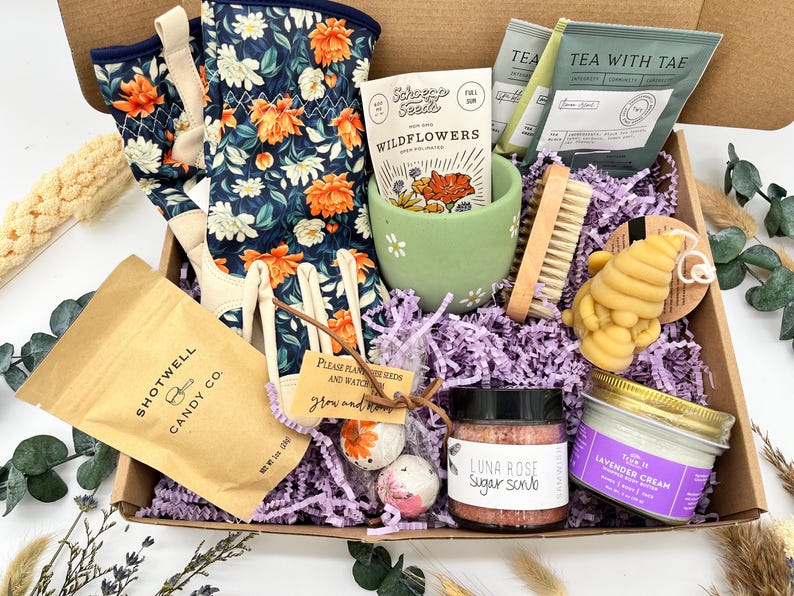 May include: A gift box filled with gardening and self-care items. Includes floral gardening gloves, wildflower seeds, tea, a sugar scrub, lavender cream, a bee-shaped candle, and candy. The box is lined with purple paper.