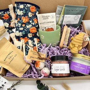 May include: A gift box filled with gardening gloves, a mug, tea, a candle, bath bombs, and a sugar scrub. The gloves have a navy blue floral pattern with orange and white flowers. The box also contains a bag of candy.