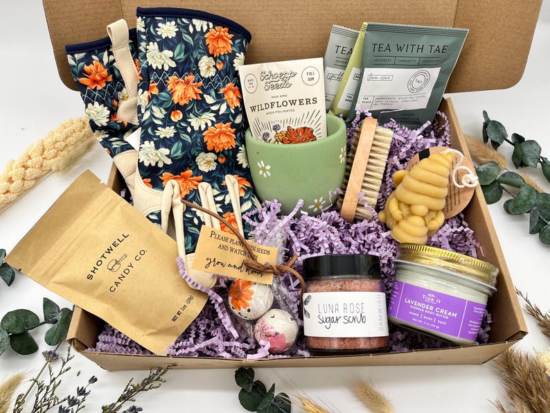 May include: A gift box filled with gardening gloves, a mug, tea, a candle, bath bombs, and a sugar scrub. The gloves have a navy blue floral pattern with orange and white flowers. The box also contains a bag of candy.