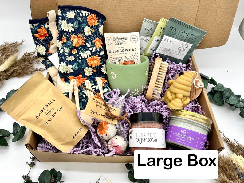 May include: A large gift box filled with gardening and self-care items. Includes floral gardening gloves, wildflower seeds, tea packets, a green ceramic cup, a wooden brush, a bee-shaped soap, sugar scrub, lavender cream, and candy. The box is labeled "Large Box".