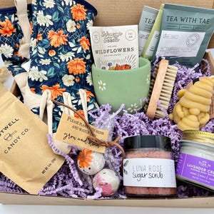 May include: A gift box filled with gardening and self-care items. Includes floral gardening gloves, wildflower seeds, tea, a sugar scrub, lavender cream, a bee-shaped candle, and candy. The box is lined with purple paper.