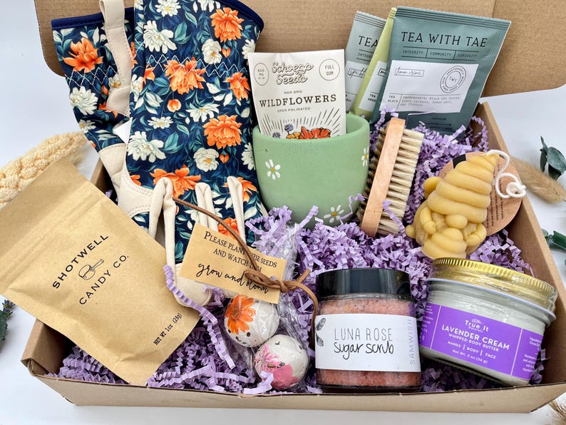 May include: A gift box filled with gardening and self-care items. Includes floral gardening gloves, wildflower seeds, tea, a sugar scrub, lavender cream, a bee-shaped candle, and candy. The box is lined with purple paper.