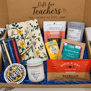 May include: A cardboard gift box labeled Gift for Teachers containing a floral notebook, three pens, a round sticker with sunflowers and the text Teacher, a candle, a bag of energy bites, a tea tin, coffee, and an Apple a Day granola bar.