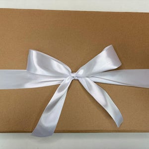 Puede incluir: Una caja rectangular de cart&oacute;n marr&oacute;n envuelta con una cinta de sat&eacute;n blanca, ancha y lisa, atada en un lazo decorativo en el centro. La caja tiene una superficie plana y lisa, y la cinta est&aacute; colocada horizontalmente en el medio.