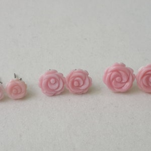 May include: Several pairs of light pink rose-shaped stud earrings. The earrings are made of a smooth material and have a detailed floral design. The earrings are displayed on a white surface.