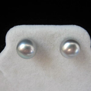 May include: A pair of round, gray pearl stud earrings. The pearls have a lustrous, iridescent sheen, reflecting light with hints of silver and blue. The earrings are displayed on a white, textured surface against a black background.
