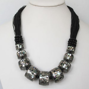May include: A statement necklace featuring large, square beads with a mosaic-like design of iridescent shell pieces. The beads are strung on black cords, with smaller black beads at the top and between the larger beads. The necklace is displayed on a white mannequin.