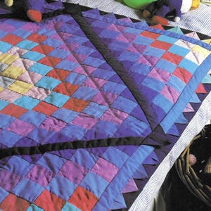 May include: A handmade quilt with a geometric diamond pattern in shades of blue, purple, red, yellow, and pink. The quilt has a black border and a scalloped edge. The quilt is likely for a baby or child.