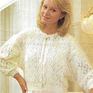 May include: A cream-coloured, open-weave cardigan featuring a diamond pattern. The long-sleeved sweater has a round neckline with a ribbon tie and a single button closure. The bottom hem and cuffs are ribbed. The cardigan is paired with a light pink skirt.