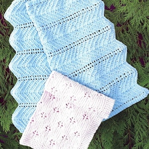 May include: Two crocheted blankets. One is light blue with a zigzag pattern and scalloped edges. The other is a smaller, square, light pink blanket with a floral design. The blankets are displayed on a bed of green foliage.
